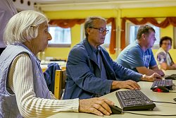 Zu sehen sind vier zum Teil ältere Personen, die an einem langen Tisch nebeneinander sitzen, vor sich jeweils eine Comutertastatur mit Maus.