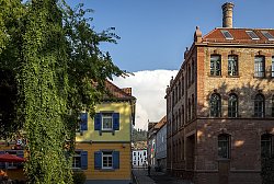 Blick auf die alte Tonofenfabrik, rechts im Bild. Es ist ein großes rotes Ziegelsteingebäude mit vielen Fensten und einem hohen runden Schornstein. Links ein gelbes Haus mit rotem Dach und blauen Fensterläden und ein großer grüner Baum. Zwischen den Häusern ist eine Gasse, an deren Ende das moderne Wohngebäude "Goldener Winkel" zu sehen ist. Über allem ein hellblauer Himmel mit einer großen weißen Wolke.
