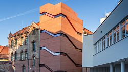 Zu sehen ist das Gebäude Stadtmuseum Tonofenfabrik in Lahr. Es beginnt auf der linken Seite mit dem alten Gebäude der ehemaligen Tonofenfabrik, ein Ziegelbau mit mit rotem Ziegeldach und rundbogenfenstern sowie einem kleinen Giebel, der nicht über die gesamte Hausbreite reicht. Rechts schließt sich ein moderner Anbau an. Er ist farblich in Ziegelrot gehalten und die Außenfassede ist gekennzeichnet durch schmale Fenster, die einem Treppenverlauf folgen.
Ganz rechts auf dem Bild ist ein Nachbargebäude mit weißer Fassade zu sehen.