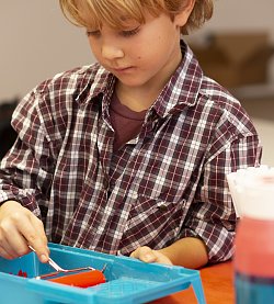 Zu sehen ist ein Junge mit blondem Haar, der sehr konzentriert mit einer Farbrolle arbeitet. Er trägt ein dunkelrot-weiß kariertes Hemd und ein dunkelrotes T-Shirt darunter. Gerade taucht er die Farbrolle in ein Gefäß mit oranger Farbe.
