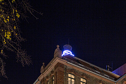 Man sieht die Gesamtansicht des Stadtmuseums. Es ist dunkel. Ein quadratischer Neonstreifen und ein runder Neonstreifen, die beide um den Kamin angeordnet sind, leuchten. Sie bildern den ersten Teil des Logos des Stadtmuseums, der für die Tonofenfabrik steht: Ein Kreis in einem Quadrat.