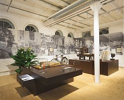 Blick in einen Raum im Stadtmuseum Lahr. Auf zwei großen braunen Ausstellungstischen sind verschiedene Exponate zu sehen, zum Teil in Glaskästen. Dahinter eine Stellwand mit großen historischen schwarz-weiß Fotografien. Eine große Grünpflanze und eine altes Fahrrad stehen zwischen Tisch und Stellwand. In der Mitte des Raums eine weiß gestrichene Stahlsäule. Der Boden ist aus hellem Holz.