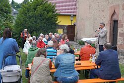 Markus Ibert mit Mikrofon an der Burgheimer Kirche, davor sitzen circa 30 Gäste auf Bierbänken.