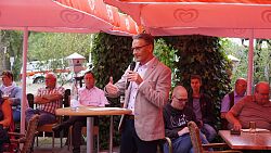 Markus Ibert mit Mikrofon am Stehtisch unter roten Sonnenschirmen, im Hintergrund sitzen Gäste