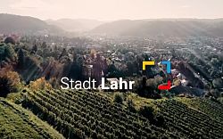 Blick über sonnige Weinberge auf die Lahrer Innenstadt. Schriftzug &#34;Stadt Lahr&#34; und das quadratische Logo mit den Farben Rot, Blau, Gelb und Schwarz.