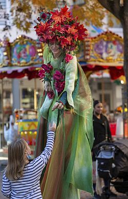 Eine Stelzenläuferin gibt einem Kind auf dem Lahrer Wochenmarkt etwas in die Hand; im Hintergrund steht ein Kinderkarussell.