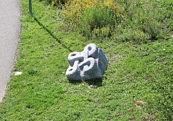 Steinskulptur in Form einer verschlungenen Schnur, auf einer Wiese liegend.