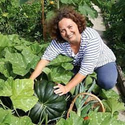 Das Bild zeigt Susanne Oswald kniend inmitten eines grünes Betes. Abstützen tut Sie sich dabei auf einem großen grünen Kürbis.