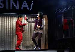 Ein Mann mit kinnlangen schwarzen Haaren und weiß angemaltem Gesicht sprüht großzügig Haarspray auf den Haaransatz eines anderen Mannes, der die Augen zusammenkneift.