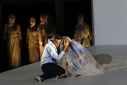 Szene aus "Die Perlenfischer". Mann und Frau hocken am Boden, im Hintergrund Menschen in weißen Gewändern.