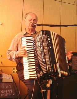 Lothar Baumann sitze mit einem brauen Hemd begleitet auf einem Hocker. Vor sich hat er sein schwarzes Akkordeon. Er hat die Augen halb geschlossen und singt in ein vor ihm stehendes Mikrofon.