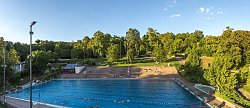Blick auf das Schwimmerbecken im Terrassenbad Lahr, dahinter die terrassenförmige Anlage mit Liegewiese und Bäumen. Im Becken schimmen wenige Menschen und auf der Anlage sind ebenfalls ereinzelt Menschen zu sehen.