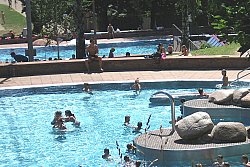 Zu sehen ist das Nichtschwimmerbecken im Terrassenbad Lahr. Dahinter sieht man einen teil des Schwimmerbeckens. rechts im Bild eine Treppe, die ins türkisblaue Wasser des Beckens führt. Im Becken sind einige Badende zu sehen. Markant erhebt sich ein halbkreisförmiges Element aus Beton im Becken mit eine Vorrichtung aus Edelstahl, aus der zu bestimmten Zeiten Wasser fließt. Auf dem Betonelement liegen große Steine.