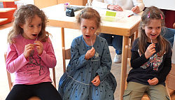 Drei Kita-Kinder sitzen auf Stühlen und reiben sich mit den Teststäbchen in den Wangen. 