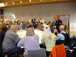 Um Tische versammelte Menschen diskutieren über das Thema "Gemeinschaft", ersichtlich an der Pinnwand im Hintergrund