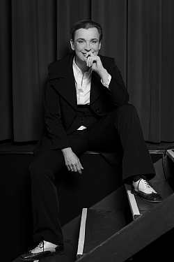 Frau in schwarzem Anzug mit Weste, weißer Bluse, sitzt auf einem Bühnenpodest und lächelt in die Kamera. Sie hat die Haare zu einem Zopf zusammengebunden und trägt schwarz-weiße Budapester-Schuhe.