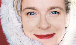 Tina Teubner trägt eine weißen Pelz um den Kopf. Ihre roten Lippen passen farblich zum Hintergrund. Ihre blauen Augen leuchten.