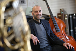 Tobias Meinen sitzt lässig mit aufgestelltem Ellenbogen in schwarzem Anzug mit grauem Pullover. Im Vordergrund verschwommen ein Tenorhorn, im Hintergrund ein Kontrabaß.