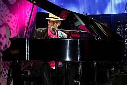 Zu sehen ist Sänger Andreas Schulte bei seiner Show &#34;Tribute to Elton John&#34;. Er sitzt am Klavier in pinkem Hemd, schwarzem Sacko und mit einem Hut. In Elton-John-Manier trägt er eine Brille mit farbigen Gläsern. 