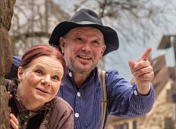 Eine Frau mit brauner Bluse, roten Haaren, die zu einem Knoten zusammengesteckt sind, und ein Mann mit blau-weiß gestreiftem Hemd, Hosenträger und Hut schauen neugierig hinter einer Mauer hervor.