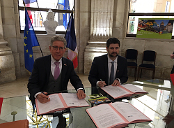 Die beiden Bürgermeister sitzen in Dole im Rathaus hinter einem Schreibtisch. Beide haben vor sich den Vertrag und schauen mit Kugelschreiber in die Hand in die Kamera. 