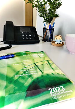 Das Programmheft der Volkshochschule Lahr auf einem Schreibtisch. Im Hintergrund ein Telefon, eine Pflanze, Stifte, eine Dekofigur und eine Tasse.