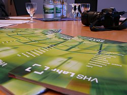 Auf einem Tisch liegen fächerförmig mehrere VHS Programmhefte. Sie sind in verschiedenen Grüntönen gehalten. Außerdem liegt auf dem Tisch ein Fotoapparat und es stehen Mineralwasserflaschen und Wassergläser im Hintergrund.