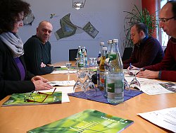 Bei der Pressekonferenz zur Vorstellung des neuen VHS Programms sitzen die Leiterin der Lahrer Volkshochschule, Carmen Wenkert, der Leiter des Amts für Außerschulische Bildung der Stadt Lahr, Tobias Meinen, mit Medienvertreter an einem Tisch zusammen. Vor ihnen liegen mehrere neue VHS Programmhefte. Zu sehen sind außerdem Gläser und Mineralwasserflaschen auf dem Tisch. Ein Medienvertreter notiert etwas auf einem Blatt Papier. Im Hintergrund ist an der Wand eine große Karte vom Landesgartenschaugelände zu sehen.