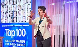 Silke Weigang steht auf einer Bühne und spricht zu ihrem Publikum. Sie gestikuliert mit den Armen. In einer Hand hält sie einen großen Filzstift. Silke Weigang hat kurzes braunes lockiges Haar und trägt eine beige Jacke, ein schwarzes Shirt und eine schwarze Hose, dazu eine große türkisfarbene Halskette und einen bunten Armreif.