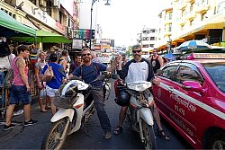 Zu sehen sind zwei Männer auf Motorrollern, die inmitten eines dichten Verkehrsgewimmels auf einer Straße stehen. Um sie herum zahlreiche Menschen und Autos.