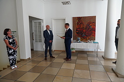 Das Foto zeigt drei Personen. Eine Person überreicht einer anderen eine Urkunde. 