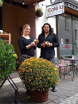 Verena Bucherer und Humberto Ovalle stehen hinter einem gelben Chrysanthemenstrauch vor ihrem Cafe, mit Schild im Hintergrund, und zeigen den Fruchtaufstrich in die Kamera. 