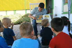 Der Pfarrer liest den Kindern vor. Er zeigt ein Bilderbuch und die Kinder lauschen ihm. 