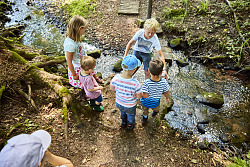 Das Bild zeigt fünf Kinder, vier Jungen und ein Mädchen, die sich bei der Überquerung über Steine des Sulzbachs helfen.
