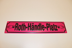 Zu sehen ist ein längliches rechteckiges Schild, das an ein Straßenschild erinnert. Darauf steht: Roth-Händle-Platz. Links und rechts des Textes ist jeweils die typische Hand von Roth-Händle abgebilet, die je nach links und rechts zeigen. Der Grundtton des Schildes ist der typische Rotton der Badischen Tabakmanufaktur Roth-Händle. Die Schrift ist in schwarz gehalten.
