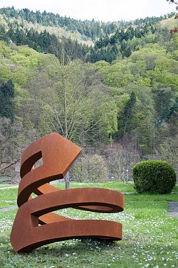 Skultpur aus Cortenstahl, wie eine Sprirale gedreht. Wiese und Wald im Hintergrund.