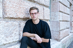 Regisseur Tom Wilmersdörffer lehnt an einer Wand mit gehauenen Steinen. Er ist ganz in dunkelbau gekleidet und trägt eine rote Brille. 