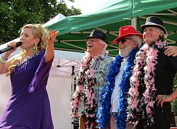 Zu sehen ist die Sängerin Catarina Conti bei ihrem Auftritt beim Wohngebietsfest Kanadaring. Sie singt in ein Mikrofon, hat langes blondes Haar und trägt ein violettes Kleid. Hinter ihr stehen drei Männer als Background Chor, die mit Hüten und bunten Federboas geschmückt sind.