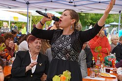 Die Sängerin Catarina Conti bei einem Auftritt beim Wohngebietsfest Kanadaring. Sie trägt ihre blonden Haare im Nacken zu einem Dutt gebunden und roten Lippenstift. Sie hat ein schwarzes Kleid mit silberner Stickerei und ein Bolerojäcken, ebenfalls schwarz, an. Sie singt in ein Mikrofon. Im Hintergrund sieht man das Publikum an Biertischen unter einem Zelt sitzen.
