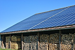 auf einer Wiese steht eine Scheune, umringt von gestapelten Holzscheiten. Auf dem dach sind mehrere Photovoltaikanlagen.