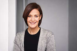 Die Autorin steht und lächelt in die Kamera. Sie hat braunes halblanges Haar und trägt ein schwarzes T-Shirt und darüber eine graue fließende Strickjacke. 