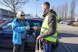 Zu sehen ist eine blonde Frau mit Sonnenbrille, die auf einem Parkplatz von Martin Stehr vom Stadtplanungsamt ein Duftherz überreicht bekommt. Die Frau trägt eine blaue Sonnenbrille und eine blaue Jacke, über dem Arm hängt ein Einkaufskorb mit weißen Punkten. Martin Stehr hat kurze blonde Haare und trägt eine gelbe Warnweste. Es ist ein sonniger Wintertag mit blauem Himmel.