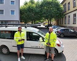 Zwei Männer in neon-gelben Jacken gekleidet, stehen vor einem weißen Dienst-Auto mit Aufdruck "Stadt Lahr". Ein Mann hält eine Fernbedienung in den Händen.