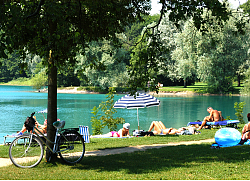 Das Bild zeigt den Waldmattensee in Kippenheimweiler. Am Ufer des von Bäumen umrahmten Baggersees liegen einige Menschen in Badebekleidung in der Sonne oder unter Sonnenschirmen. An einem Baum lehnt ein Fahrrad.