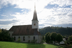 Das Bild zeigt die Kirche Mariä Heimsuchung in Kuhbach im Gegenlicht. Der Spitze Turm mit dem langen Kirchenschiff dahinter steht zwischen Bäumen an einer Wiese. Im Hintergrund sind Schwarzwaldberge sowie rechts Häuser zu sehen.