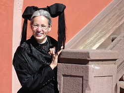 Das Bild zeigt Stadtführerin Annemarie Friedrich-Kirn mit silberner Nickelbrille und grauen, zusammengebundenen Haaren in schwarzer Tracht mit einer großen schwarzen Schleife auf dem Kopf an das Treppengeländer des Alten Rathauses gelehnt.