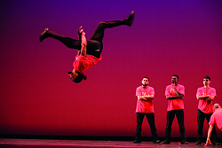 Ein Tänzer schlägt im Vordergrund einen Salto, drei Tänzer schauen im Hintergrund zu. Alle tragen schwarze Hosen und ein rotes T-Shirt. Die Bühne ist in rot gehalten.
