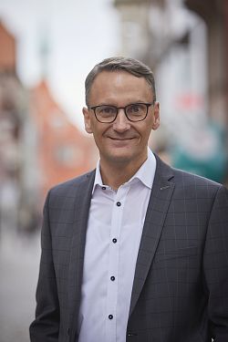 Foto von Oberbürgermeister Markus Ibert, mit weißem Hemd und grauem Sakko, im Hintergrund verschwommen Häuser