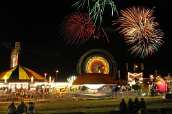 Das Bild zeigt einen Rummelplatz in Belleville bei Nacht. In der Mitte ist ein bunt leuchtendes Riesenrad zu erkennen, rings herum weitere hell erleuchtete Karussells und Fahrgeschäfte. Am Himmel ist ein Feuerwerk zu sehen.