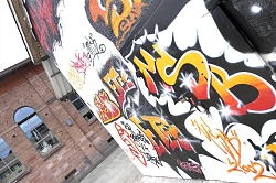 Das Bild zeigt den Schlachthof zwischen gestern und heute. Im Hintergrund ist ein altes Backsteingebäude zu erkennen, rechts eine moderne Graffiti-Wand.